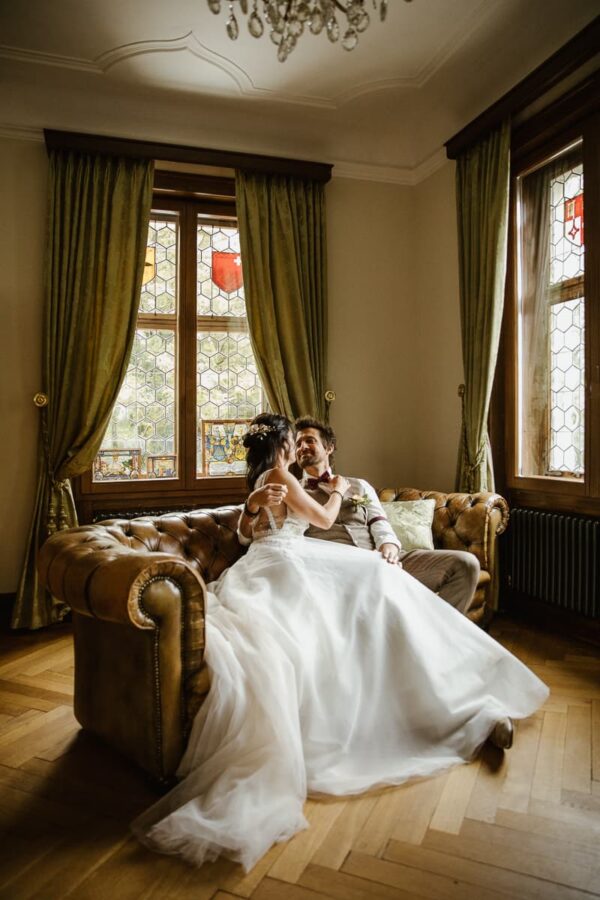 Brautpaar sitzt gemeinsam auf dem Chesterfield Sofa im Kaminzimmer des Schlössli Utenberg und lacht sich an.