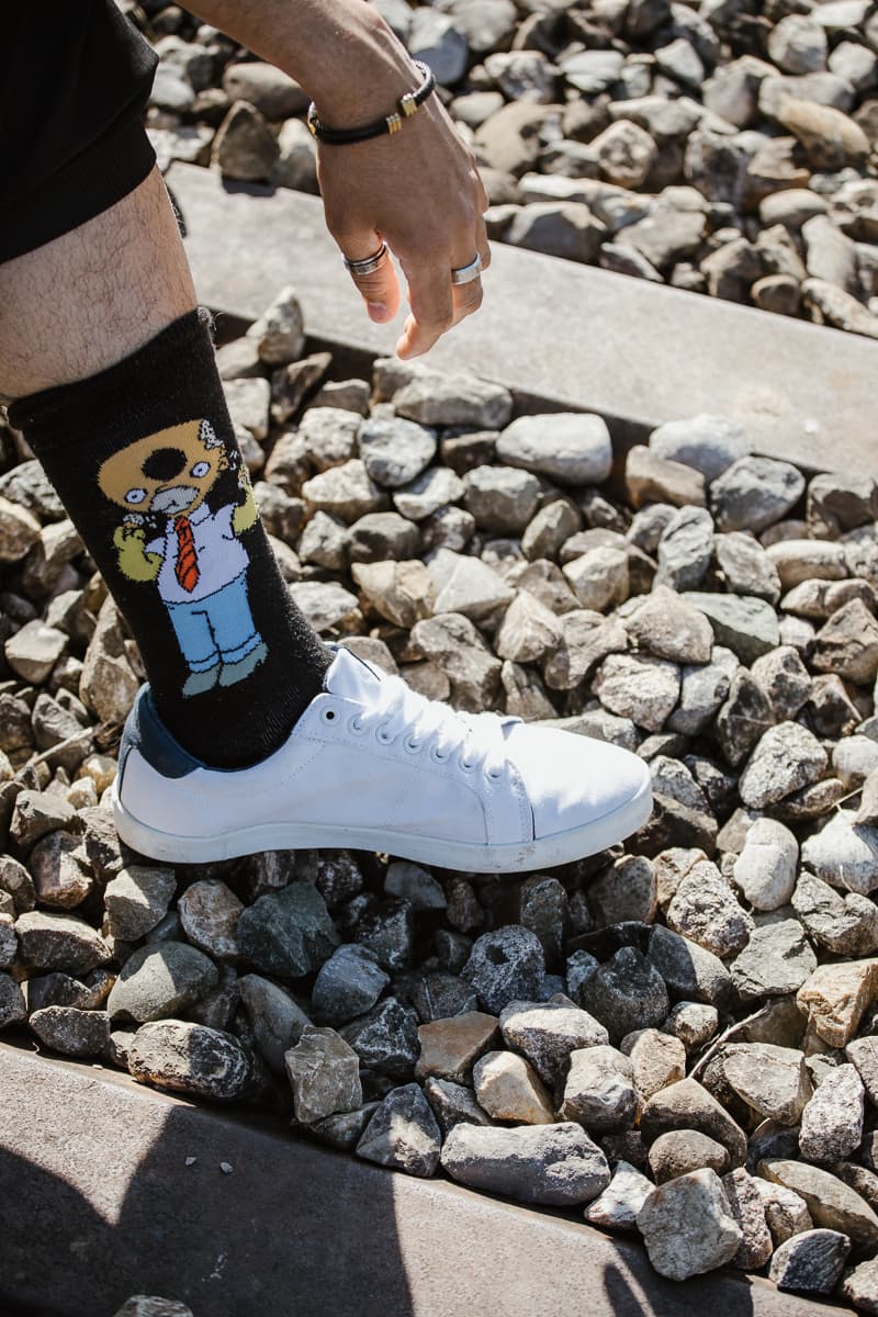 Detailaufnahme einer schwarzen Socke mit Comic-Motiv und weissem Sneaker auf Kiesuntergrund, Hand mit Ringen im Bild