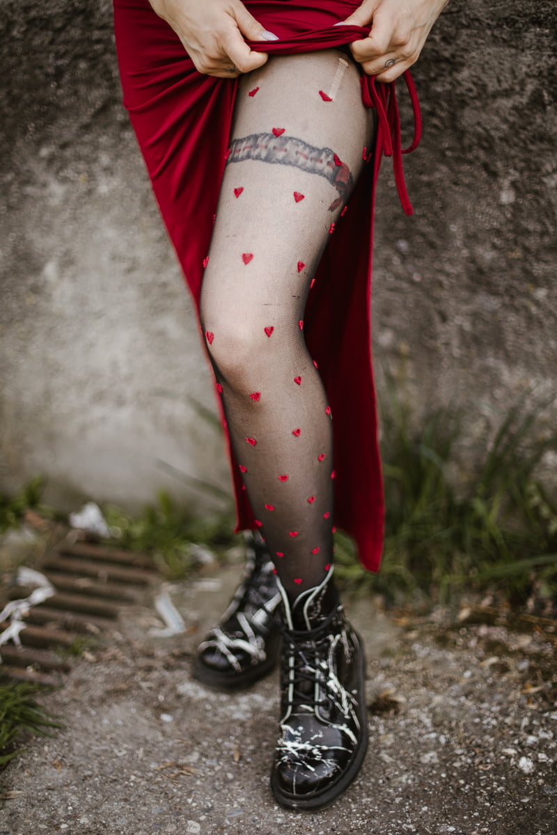 Detailaufnahme eines Beins mit Strumpfhose im Herzchenmuster unter gerafftem rotem Kleid, schwarze Boots