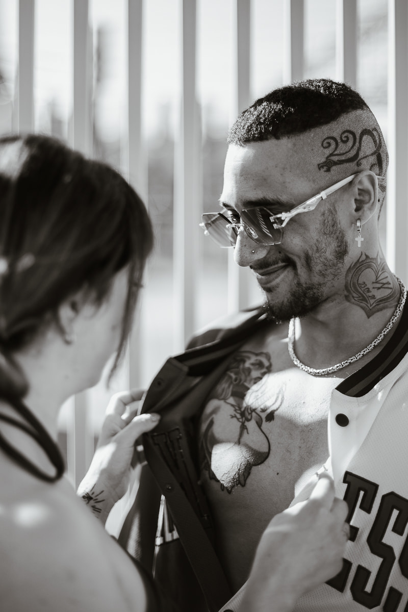 Schwarzweiss-Foto: Frau oeffnet laechelnd das Trikot ihres Mannes, sein Brust-Tattoo wird sichtbar